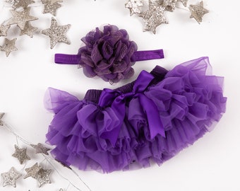 May include: Purple tulle tutu with a matching purple satin bow and a purple flower headband. The tutu is made of multiple layers of tulle and has a ruffled edge. The headband is adorned with a large, layered flower.