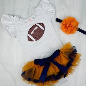 May include: A white bodysuit with a brown glitter football design, a navy blue and orange tulle tutu with a navy blue bow, and an orange flower headband.
