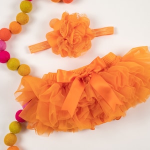 May include: Orange tulle baby bloomers with a matching satin bow and a flower headband. The headband has a large orange flower with a white lace center.