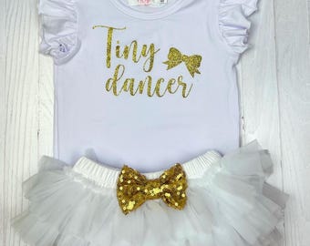 May include: White baby onesie with gold glitter text that says "Tiny dancer" and a gold glitter bow. The onesie is paired with a white tulle tutu with a gold glitter bow.