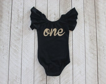 May include: Black bodysuit with gold glitter lettering that says "one". The bodysuit has short, ruffled sleeves.