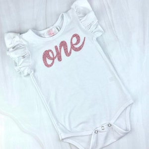 May include: White baby onesie with pink glitter lettering that says "one". The onesie has ruffled sleeves.