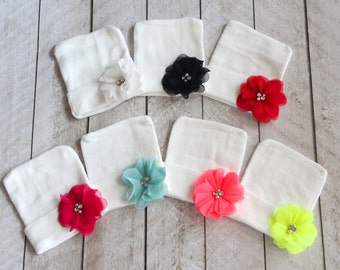 May include: Seven white baby hats with different colored fabric flowers attached. The flowers have rhinestones in the center. The colors of the flowers are red, pink, turquoise, yellow, black, and white.