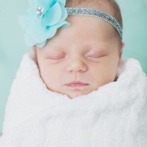 May include: A sleeping baby girl wearing a white headband with a light blue flower on top. The baby is wrapped in a white blanket.