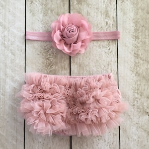 May include: A pink tulle baby bloomers with a matching pink flower headband. The bloomers are made of multiple layers of tulle and have a ruffled edge. The headband has a single large pink flower with a centre.
