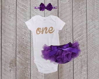 May include: A white onesie with gold glitter lettering that says "one". The onesie is paired with a purple tulle tutu and a purple bow headband.