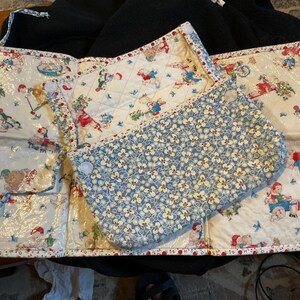 May include: A quilted folding mat featuring a white background with a repeating pattern of children playing with toys and a blue floral print flap. The edges are finished with a red and white patterned trim, and the mat includes white hook-and-loop fasteners.