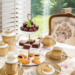 Puede incluir: Una escena de fiesta de té con una bandeja de servicio de tres niveles con cerezas, brownies y pasteles. Tazas de té, platillos y una tetera doradas dispuestas sobre un mantel floral. Una silla decorativa está en el fondo.