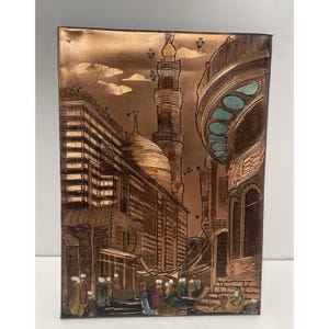 May include: A copper-toned metal wall art piece depicting a Middle Eastern cityscape. The artwork features a mosque with a tall minaret, domes, and detailed architectural elements. Figures of people are visible in the streets, and the sky has cloud details.