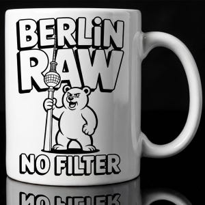 Pu&ograve; includere: Tazza in ceramica bianca con la scritta "BERLIN RAW NO FILTER" in lettere nere. Un orso dei cartoni animati che tiene una struttura alta &egrave; al centro. La tazza ha un manico curvo.