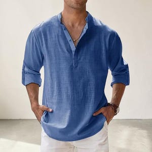 May include: A blue long-sleeved shirt with a mandarin collar and a button-down placket. The shirt has rolled-up sleeves and is made of a lightweight fabric. The model is wearing a watch and a necklace.