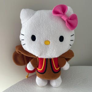 May include: A soft Hello Kitty toy with a white body, black eyes, and a yellow nose. It has a pink bow, a brown cape, and a red vest with yellow trim. The doll is standing on a white surface.