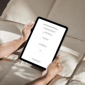 Puede incluir: Una tableta negra que muestra un planificador diario titulado "Bare Minimum Daily Planner". La pantalla muestra una lista de tareas y avisos. La tableta la sostiene una persona sobre una superficie crema acolchada.