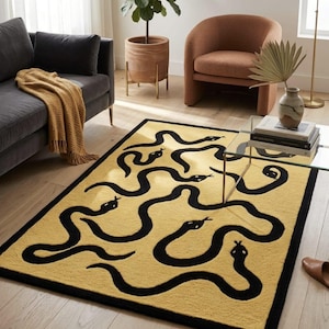 Puede incluir: Una alfombra rectangular con un fondo amarillo mostaza y un patrón repetitivo de serpientes negras. La alfombra tiene un borde negro grueso que enmarca todo el diseño, creando un contraste marcado entre los colores amarillo y negro.