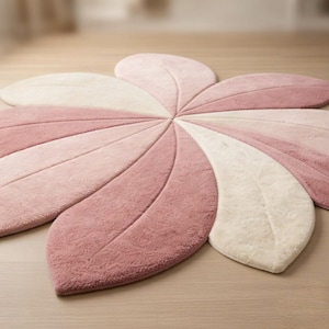 May include: A flower-shaped rug with six petals in varying shades of pink and cream. The rug is made of a soft, plush material and has a detailed leaf-like pattern. The rug is placed on a light-colored wooden floor.