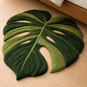 Puede incluir: Una alfombra verde con forma de hoja de monstera. La alfombra presenta diferentes tonos de verde, creando una apariencia texturizada. La alfombra está colocada sobre un suelo de madera, cerca de una cama.