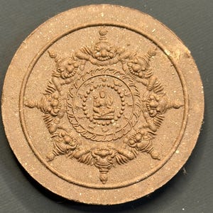 Puede incluir: Medallón redondo y marrón en relieve con una figura central sentada rodeada de un diseño ornamentado. El borde exterior del medallón tiene un aspecto texturizado, y el diseño sugiere un tema religioso o espiritual.