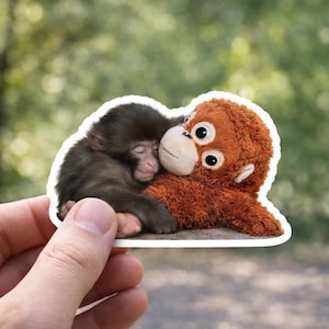 May include: A sticker featuring a baby monkey snuggling with a plush orangutan. The baby monkey has dark brown fur and is sleeping. The plush toy is orange with large, round eyes and a smiling face. The sticker is held by a hand against a blurred green background.