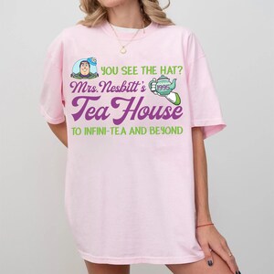 K&ouml;nnte beinhalten: Ein hellrosa T-Shirt mit einem Grafikdesign. Das Design enth&auml;lt den Text "YOU SEE THE HAT?" und "Mrs. Nesbitt's Tea House" in Lila und Gr&uuml;n, zusammen mit einem Bild von Buzz Lightyear und einer Teekanne. Der Satz "TO INFINI-TEA AND BEYOND" ist ebenfalls auf dem T-Shirt aufgedruckt.