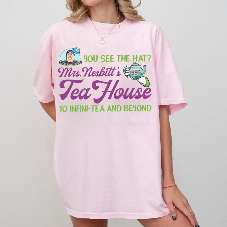 K&ouml;nnte beinhalten: Ein hellrosa T-Shirt mit einem Grafikdesign. Das Design enth&auml;lt den Text "YOU SEE THE HAT?" und "Mrs. Nesbitt's Tea House" in Lila und Gr&uuml;n, zusammen mit einem Bild von Buzz Lightyear und einer Teekanne. Der Satz "TO INFINI-TEA AND BEYOND" ist ebenfalls auf dem T-Shirt aufgedruckt.