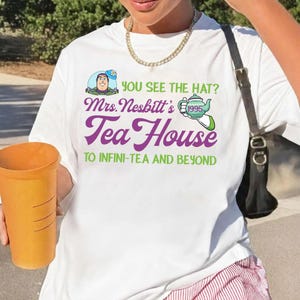 K&ouml;nnte beinhalten: Wei&szlig;es T-Shirt mit einem grafischen Design mit dem Text "YOU SEE THE HAT? Mrs. Nesbitt's Tea House TO INFINI-TEA AND BEYOND." Das Design enth&auml;lt ein Cartoon-Bild einer Figur und eine Teekanne. Die Person h&auml;lt einen gro&szlig;en orangefarbenen Becher.