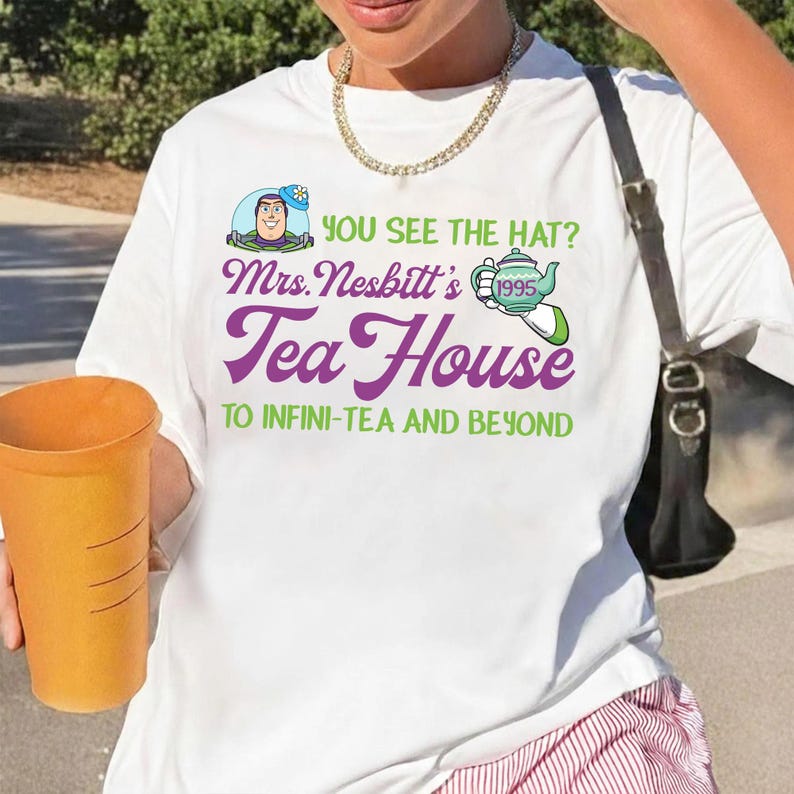 K&ouml;nnte beinhalten: Wei&szlig;es T-Shirt mit einem grafischen Design mit dem Text "YOU SEE THE HAT? Mrs. Nesbitt's Tea House TO INFINI-TEA AND BEYOND." Das Design enth&auml;lt ein Cartoon-Bild einer Figur und eine Teekanne. Die Person h&auml;lt einen gro&szlig;en orangefarbenen Becher.