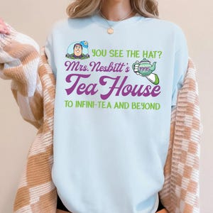 K&ouml;nnte beinhalten: Hellblaues T-Shirt mit dem Text "YOU SEE THE HAT? Mrs. Nesbitt's Tea House TO INFINI-TEA AND BEYOND". Die Grafik zeigt eine Zeichentrickfigur, eine Teekanne und das Jahr 1995. Das Shirt hat einen Rundhalsausschnitt.