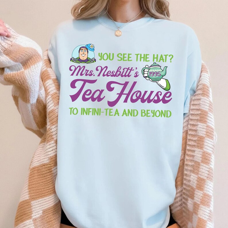 K&ouml;nnte beinhalten: Hellblaues T-Shirt mit dem Text "YOU SEE THE HAT? Mrs. Nesbitt's Tea House TO INFINI-TEA AND BEYOND". Die Grafik zeigt eine Zeichentrickfigur, eine Teekanne und das Jahr 1995. Das Shirt hat einen Rundhalsausschnitt.