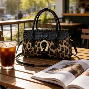 Peut inclure: Un sac à main noir et léopard avec un fermoir en forme de fer à cheval doré. Le sac a une poignée supérieure et une bandoulière amovible. Le sac est posé sur une table en bois à côté d'un verre de café glacé et d'un magazine ouvert.