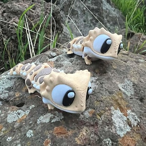 May include: Two toy lizard figures in beige and brown, featuring large, round, blue and black eyes, sit on a large rock. The lizards have a textured design and are placed outside, with green grass visible in the background.