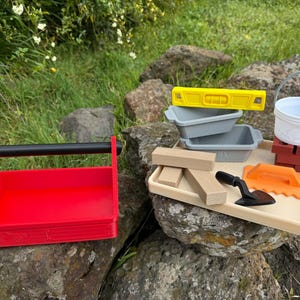 May include: A red plastic tool caddy with a black handle sits beside a beige tray containing a yellow level, two grey containers, a white bucket, three wooden blocks, a black trowel, an orange notched float, and a small red brick structure.