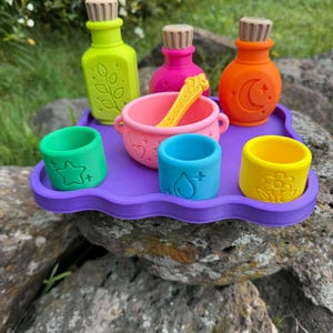 May include: A set of colourful silicone potion-making toys on a purple tray. Includes a lime green bottle with a leaf, a pink bottle, an orange bottle with a moon, a pink cauldron with a yellow spoon, and three small cups featuring star, water drop, and flower icons.
