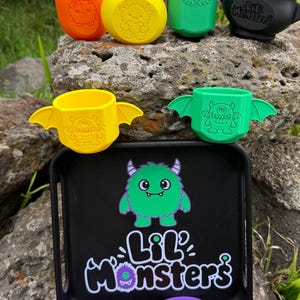 May include: A collection of plastic items including orange, yellow, and green bottles with monster engravings, a black cauldron, two winged monster cups, a black tray with a green monster graphic and the text "LiL Monsters", a purple spoon, and an orange cube.