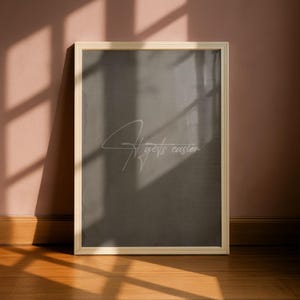 Op de afbeelding: Een ingelijste print met de tekst "It gets easier" in een cursief lettertype. De print heeft een donkergrijze achtergrond en een licht houten frame. De print leunt tegen een roze muur en een houten vloer.