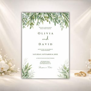 May include: A wedding invitation with a floral design. The invitation features the names Olivia and David, with the date and time of the wedding. Green leaves and olive branches adorn the top and bottom of the invitation. A lit candle and wedding rings are in the background.