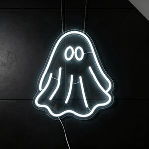 May include: A white neon ghost sign with two round eyes and a wavy bottom edge, hanging against a black background. The sign is suspended by two silver chains, and a white cord extends from the bottom.