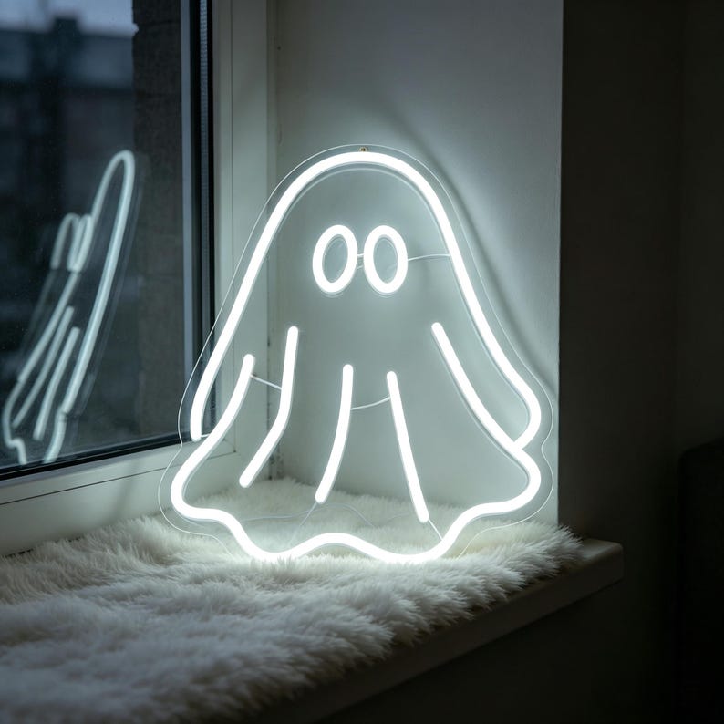 May include: A white neon ghost sign with two round eyes and vertical lines, mounted on a window sill. The neon light is bright and creates a spooky, yet playful, ambiance. The ghost is a fun decoration for Halloween or any time of year.