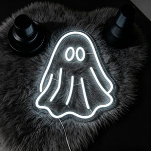 May include: A white neon ghost sign with two oval eyes and a wavy bottom edge. The ghost is lit up against a dark grey, textured background. Two black objects are on either side of the ghost.
