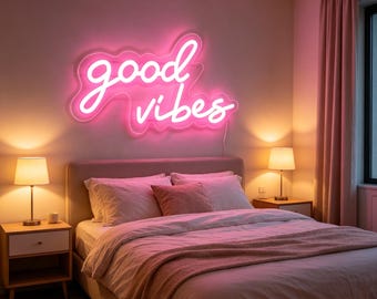 May include: A bedroom scene featuring a pink neon sign that reads "good vibes" above a bed. The bed has white linens, a pink blanket, and pillows. Two bedside tables with lamps flank the bed, and pink curtains are visible.