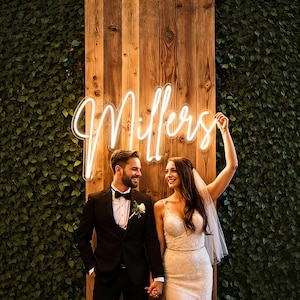 May include: A couple stands in front of a wooden wall and a green wall of leaves. A neon sign that says "Millers" is above them. The woman is wearing a white wedding dress and veil. The man is wearing a black tuxedo.
