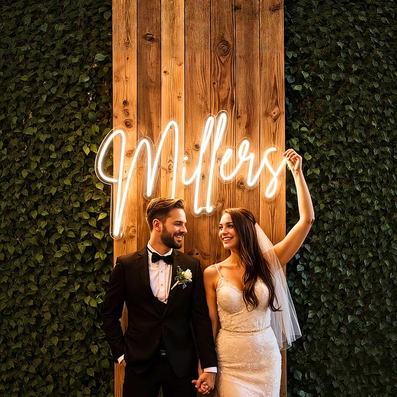 May include: A couple stands in front of a wooden wall and a green wall of leaves. A neon sign that says "Millers" is above them. The woman is wearing a white wedding dress and veil. The man is wearing a black tuxedo.