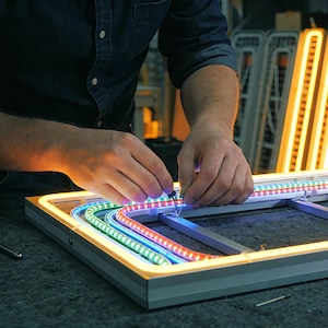May include: A person working on a neon sign. The sign is rectangular with a silver frame and colorful LED lights. The lights are red, green, and blue. The background shows other neon signs and tools.
