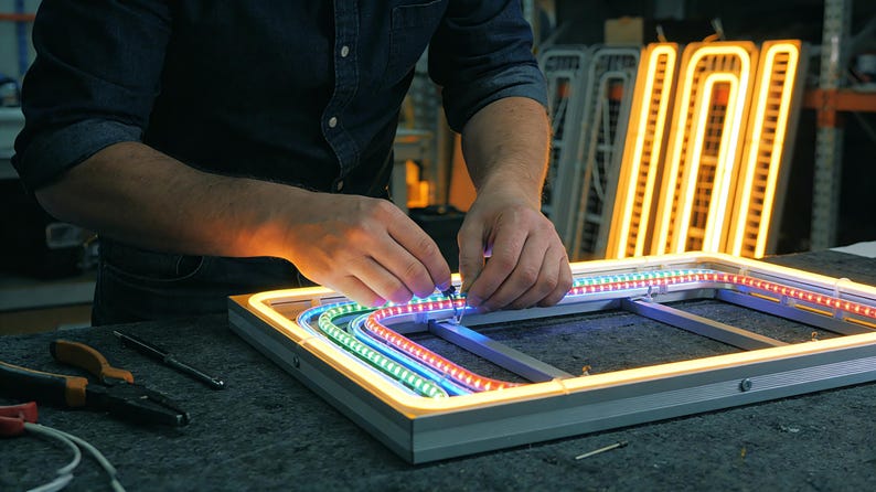 May include: A person working on a neon sign. The sign is rectangular with a silver frame and colorful LED lights. The lights are red, green, and blue. The background shows other neon signs and tools.