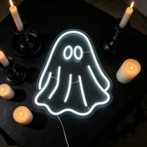 May include: A white neon ghost sign with two round eyes and a wavy bottom edge. The sign is on a black surface, surrounded by lit candles in black candle holders and several free-standing candles, creating a spooky Halloween theme.