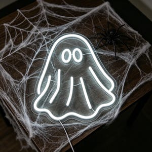 May include: A white neon ghost sign with two oval eyes and three vertical lines, resting on a wooden surface. A white spider web and a black plastic spider are also present, creating a Halloween theme.