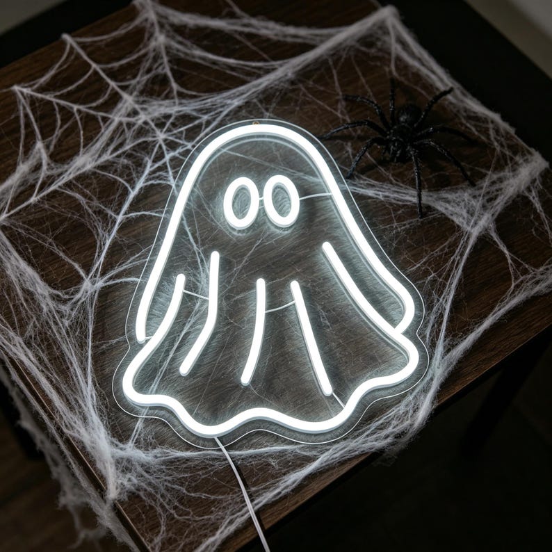 May include: A white neon ghost sign with two oval eyes and three vertical lines, resting on a wooden surface. A white spider web and a black plastic spider are also present, creating a Halloween theme.