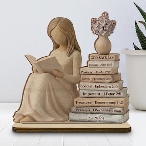 Puede incluir: Figura de madera de una mujer leyendo un libro, con una pila de libros a su lado. Los libros tienen inscripciones como "Dios dice que soy" y "Elegido". Un pequeño jarrón con flores se encuentra encima de los libros. La base es de madera.