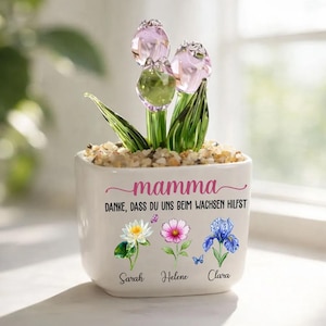 Pode incluir: Um vaso de cerâmica branca com esculturas de flores de vidro. O vaso tem a palavra "mamma" em escrita rosa, com o texto "DANKE, DASS DU UNS BEIM WACHSEN HILFST" abaixo. Ilustrações florais e nomes também estão no vaso.