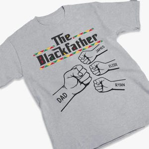 May include: A gray t-shirt with the text "The Blackfather" in black, red, green, and yellow. Below the text are four fists with the names "DAD", "JAMES", "ELISE", and "RYAN" printed on the shirt.