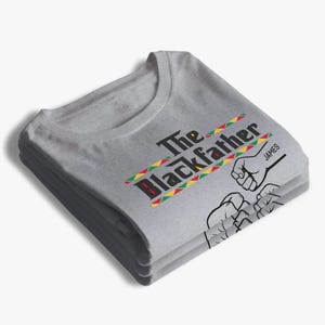 May include: A folded gray t-shirt with the text "The Blackfather" in black and red, with a colorful stripe design. The shirt also features a graphic of two fists bumping and the name "JAMES".